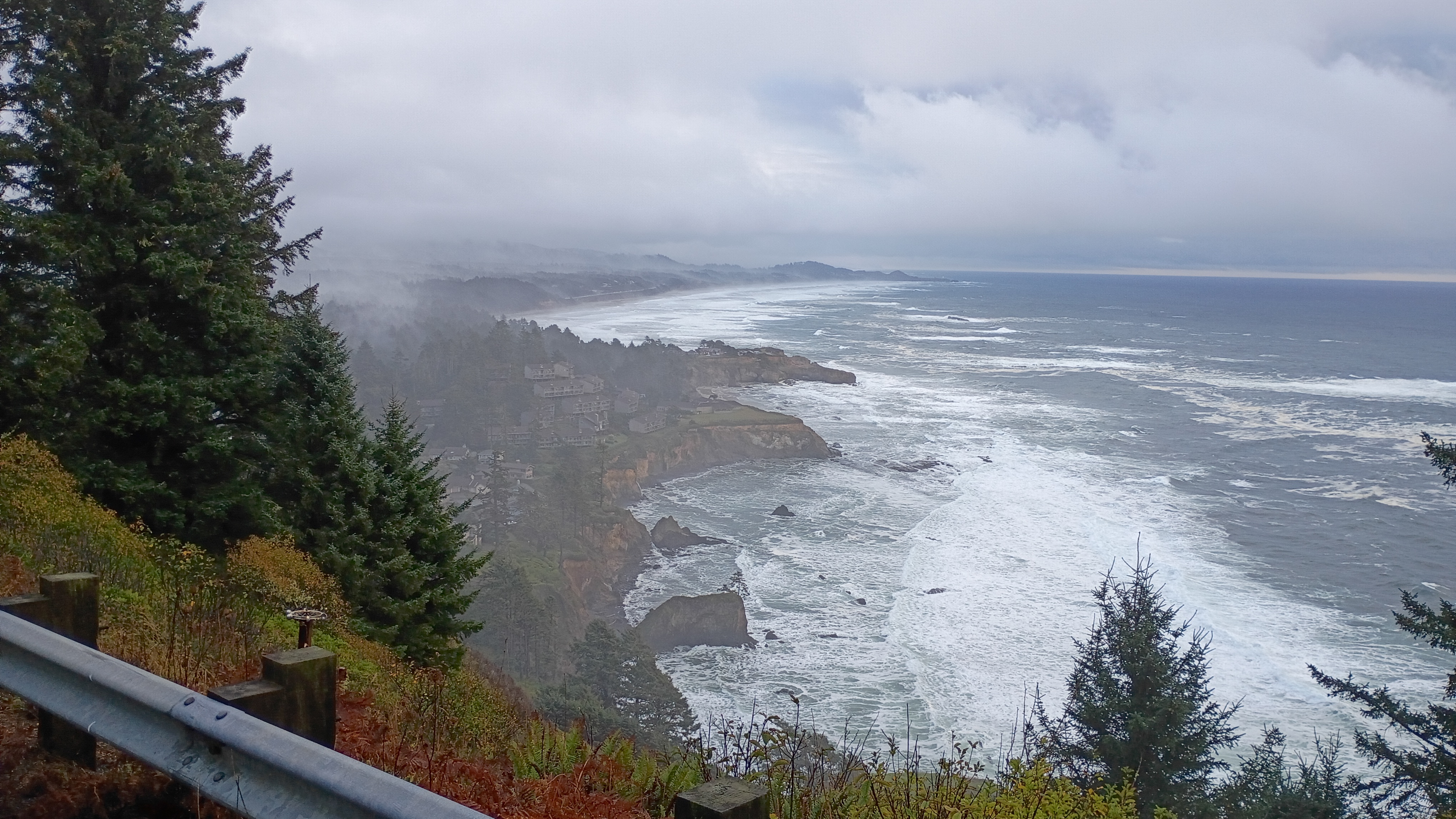 Overcast foggy day looking down on the Pacific Ocean from hundreds of feet up. Big ocean swells are crashing against rock cliffs. Houses sit perched on the edge of the cliffs. All is ringed in fog and mist and pounding surf.