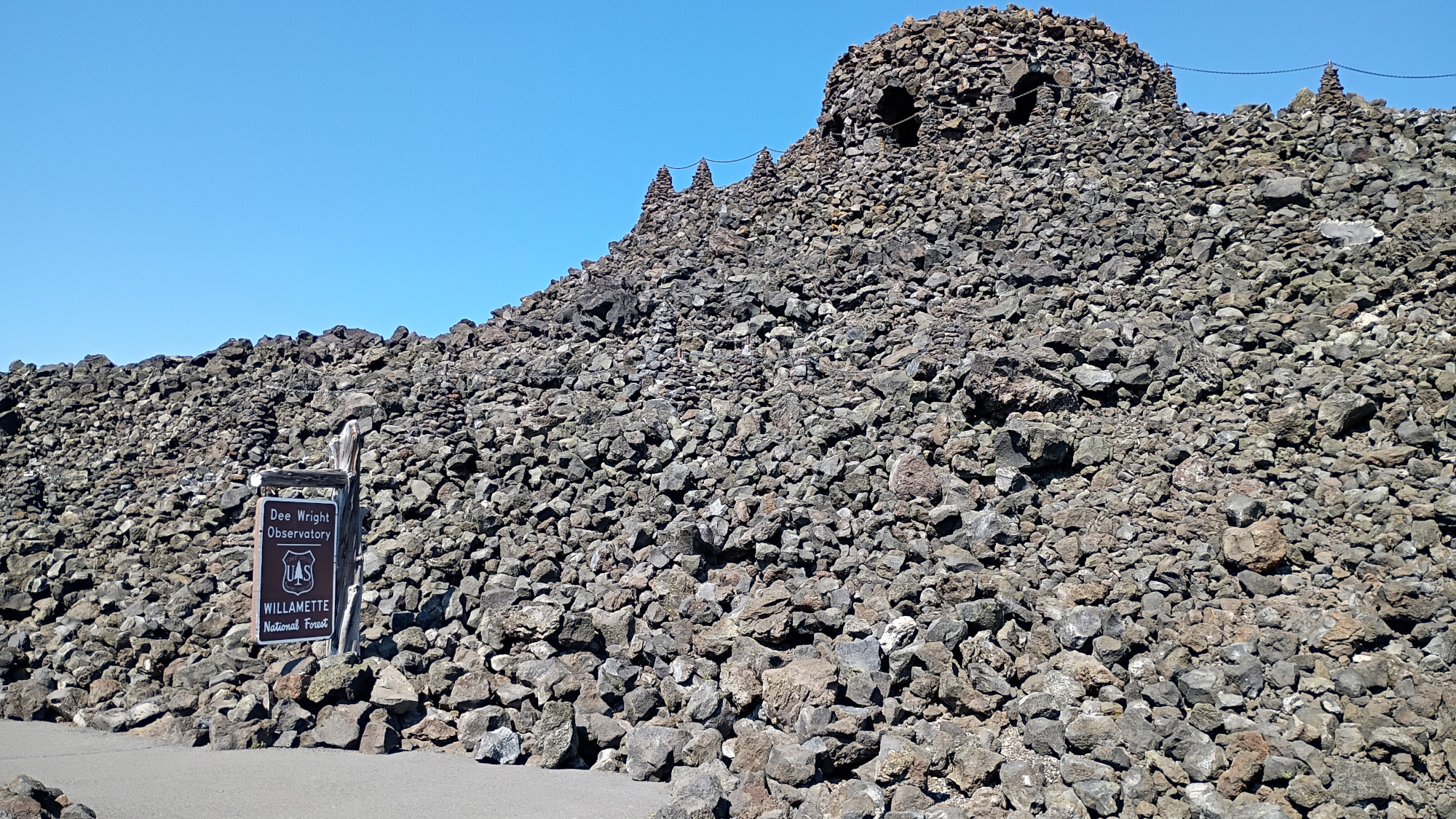 Stacked lava flow with a turret on top. Blue sky. Wooden sign read Dee Wright Observatory.