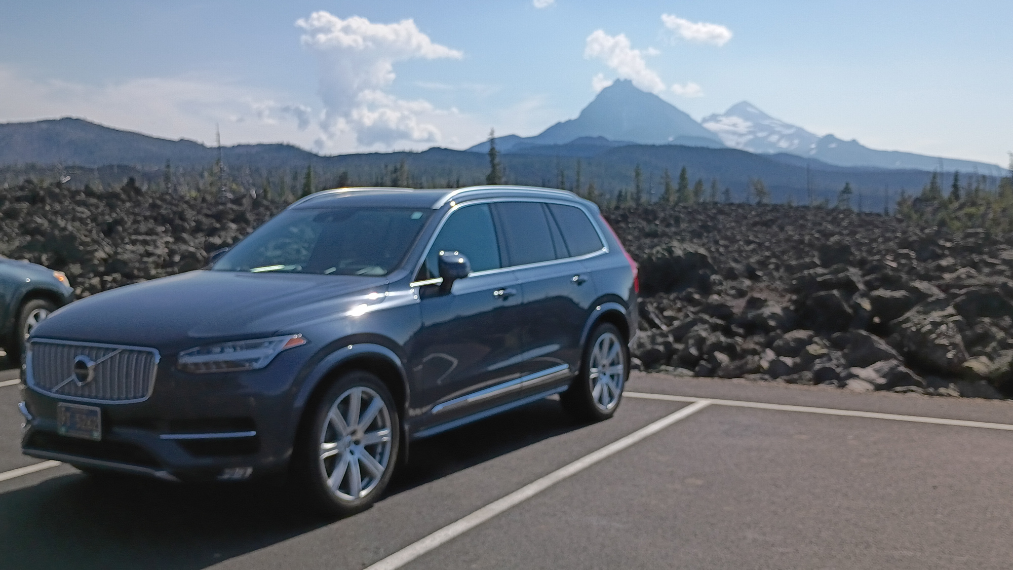 Blue XC90 in a parking space with a rocky lava field behind it and two mountain tops in the background
