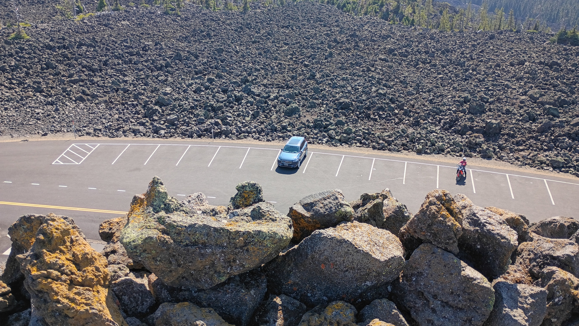 Picture from the top of the observatory looking down toward one of two parking lots. There is a blue XC90 in one parking space and a motorcycle another. Behind them is a very large lava rock field.