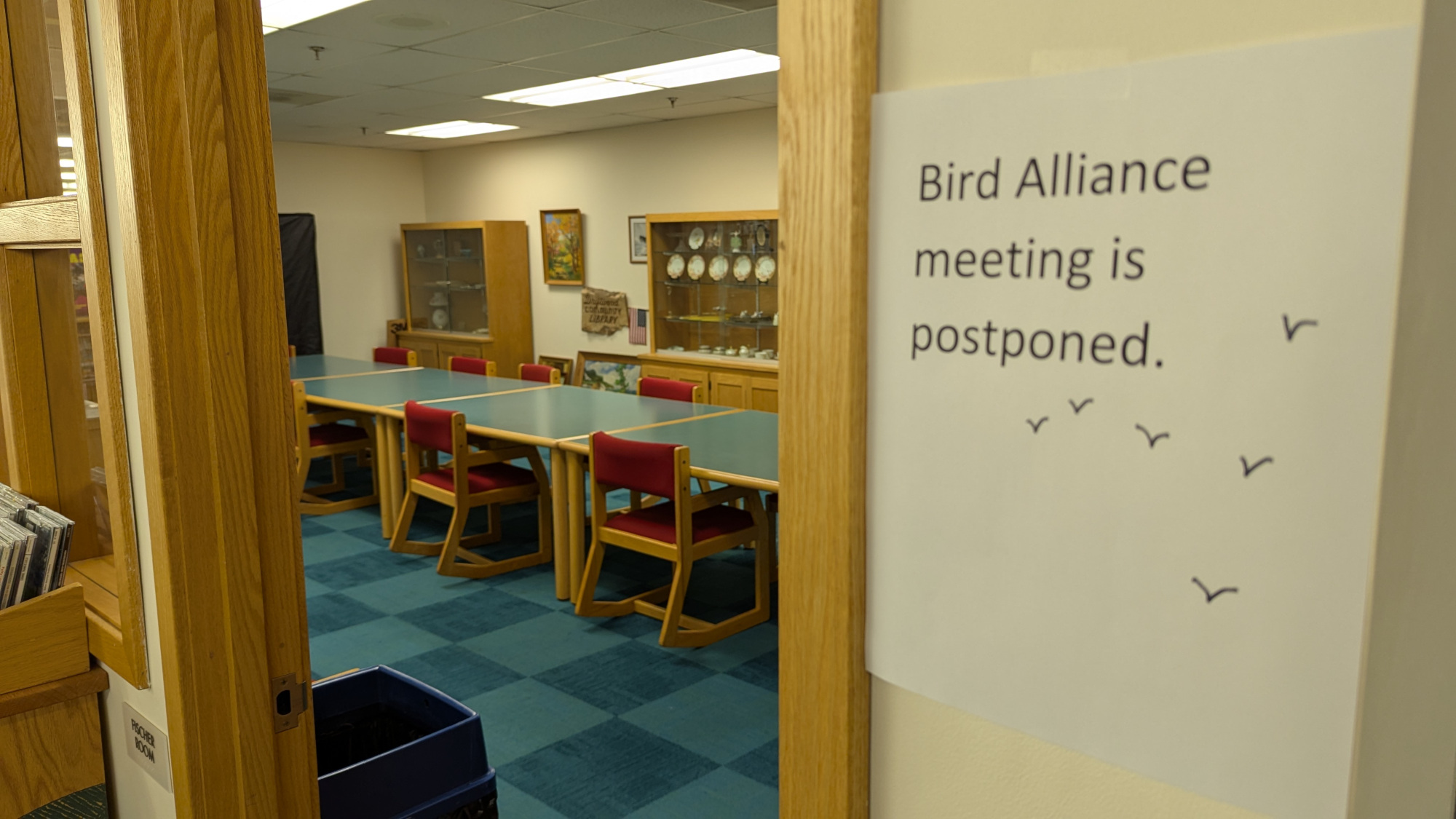 Looking into an open doorway of library meeting room. Long conference table with chairs around it. Empty. On the wall next to the doorway reads a sign stating Bird Alliance meeting is postponed. Underneath the typed message are half a dozen bird silhouettes.