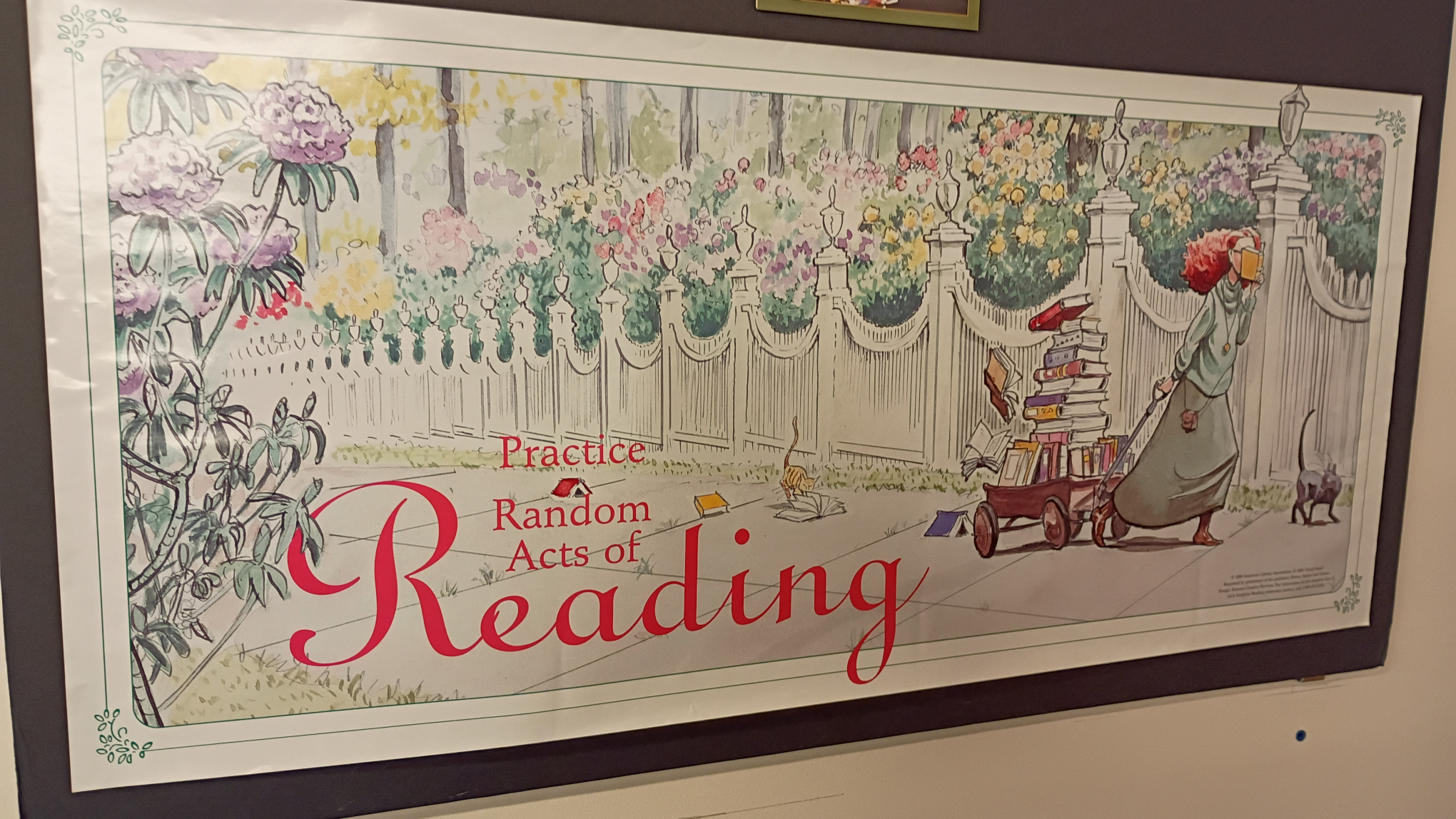 Hung print of a woman with long, red hair pulling a red wagon filled with books. There is a double sidewalk with a fancy white picket fence. Behind the fence are hundreds of tall flowers threatening to overflow the fence. A couple of the books have fallen from the wagon. One book, having fallen face up, is being read by a little brown cat. A little gray cat is in front of the woman, looking back. Text on the print reads Practice Random Acts of Reading.