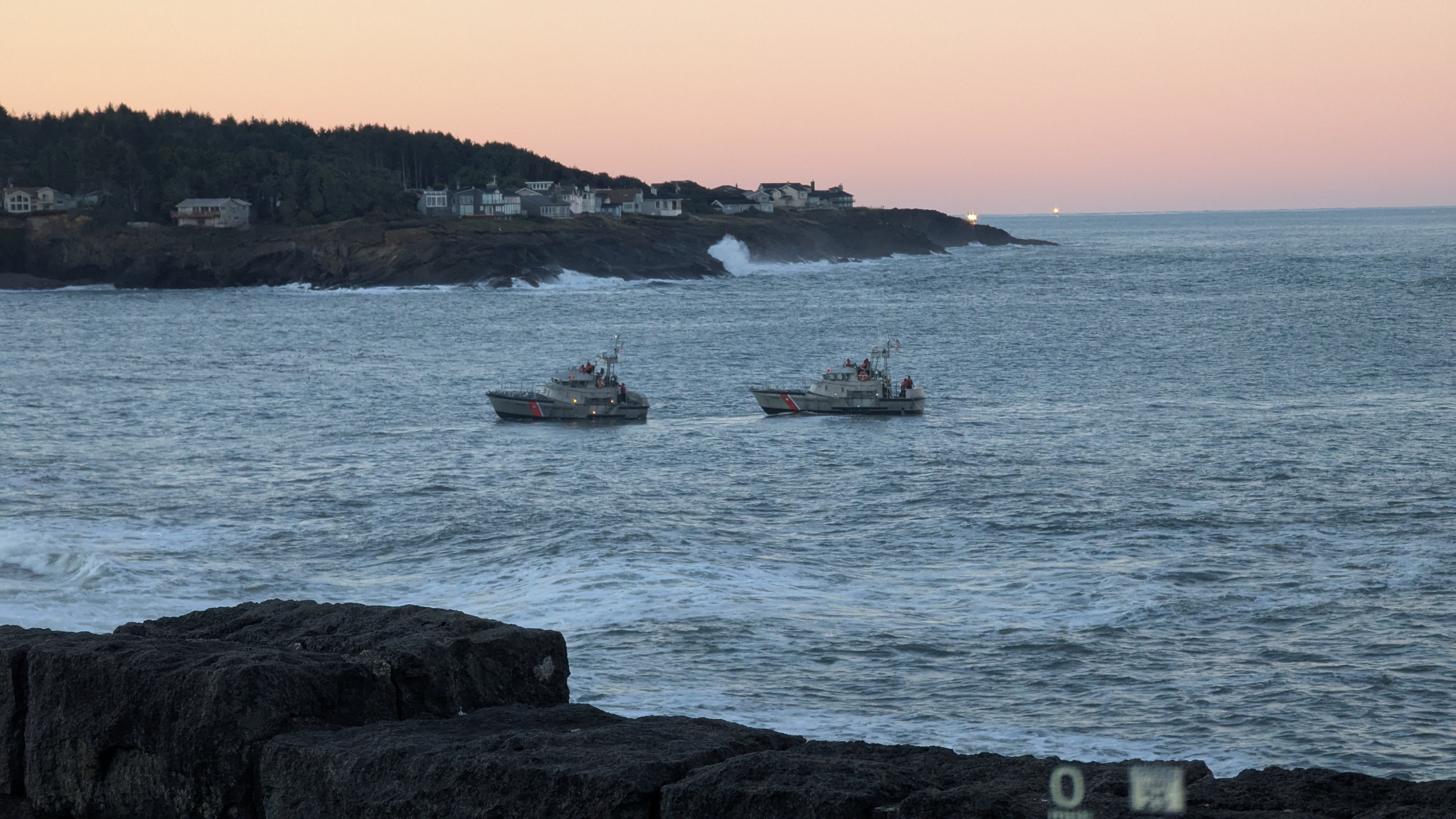 Early morning. Sky is painted Easter colors. Sea is slate blue and fairly calm. One Coast Guard ship is towing another Coast Guard ship into port. Even though it’s calm, waves are crashing onto the volcanic rock shore.