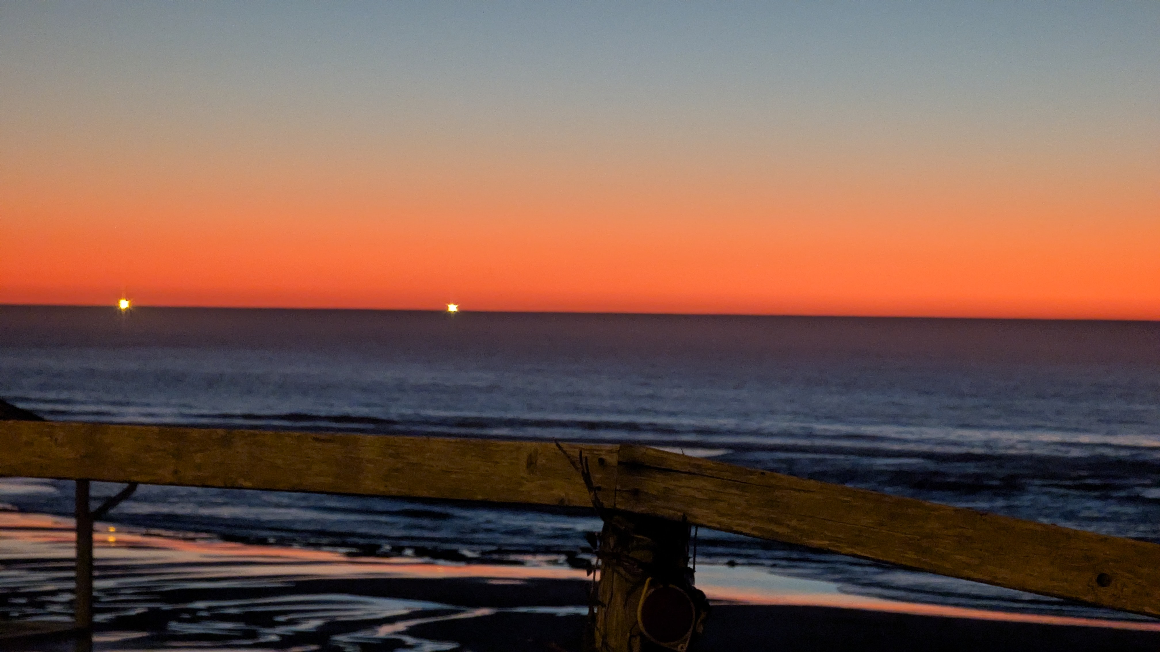 After sunset. On a bluff overlooking the Pacific Ocean. Wooden Fence in foreground. Pacific Ocean is a dark slate blue. Sky is watercolor orange fading to watercolor blue. Two fishing vessels shine bright lights on the horizon.