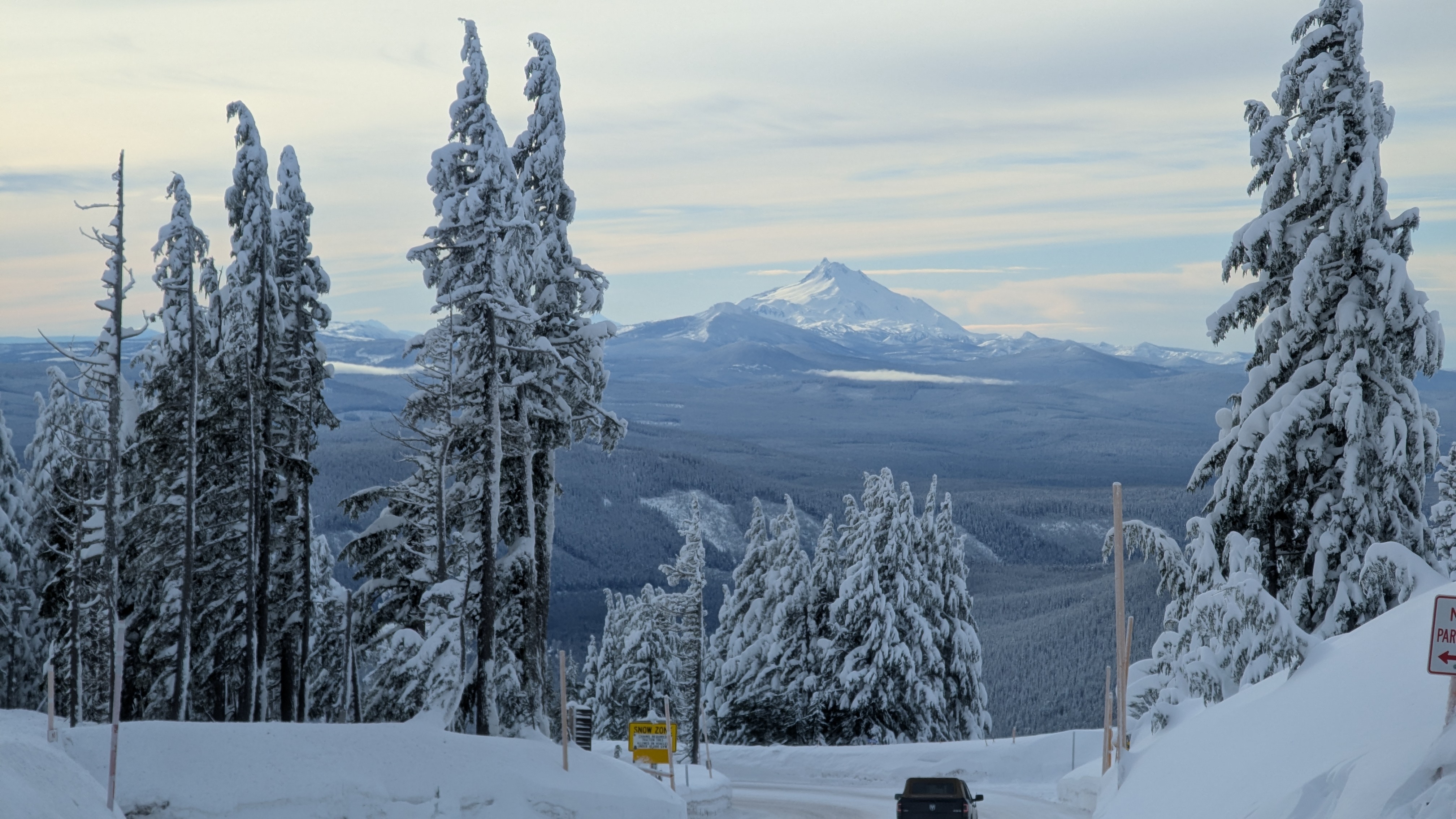 Daytime. Snowy mountain road. Heading down the mountain. A black pickup is ahead in the distance. The snow bunched up around the road is a good fifteen feet high. Conifers are covered in heavy snow. My. Jefferson sits in the distance.