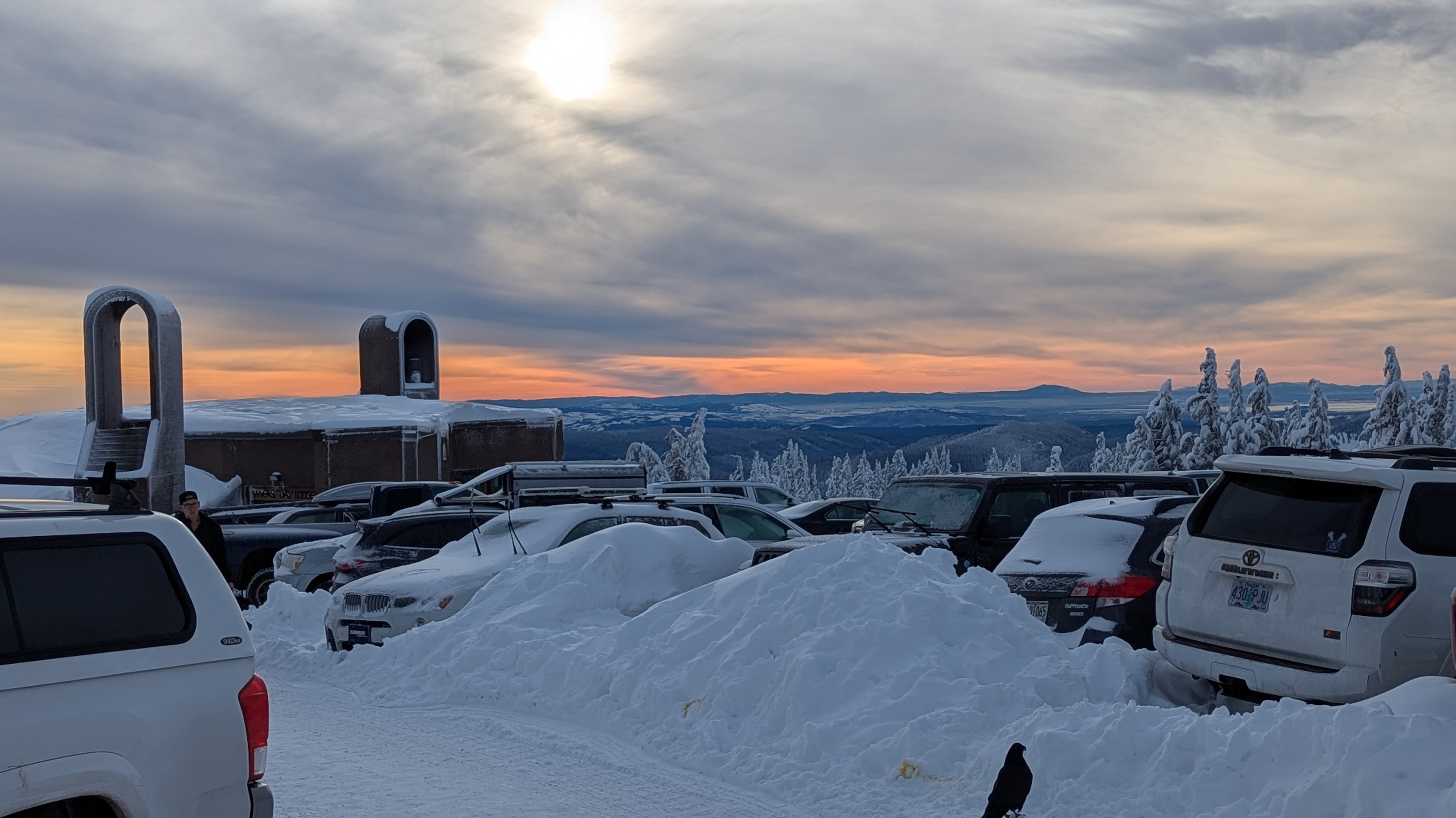 Daytime. Snowy mountain. Cars in foreground buried under snow. Sky is beautiful yellow, orange, and steel blue gray. Sunlight barely shines through the clouds.