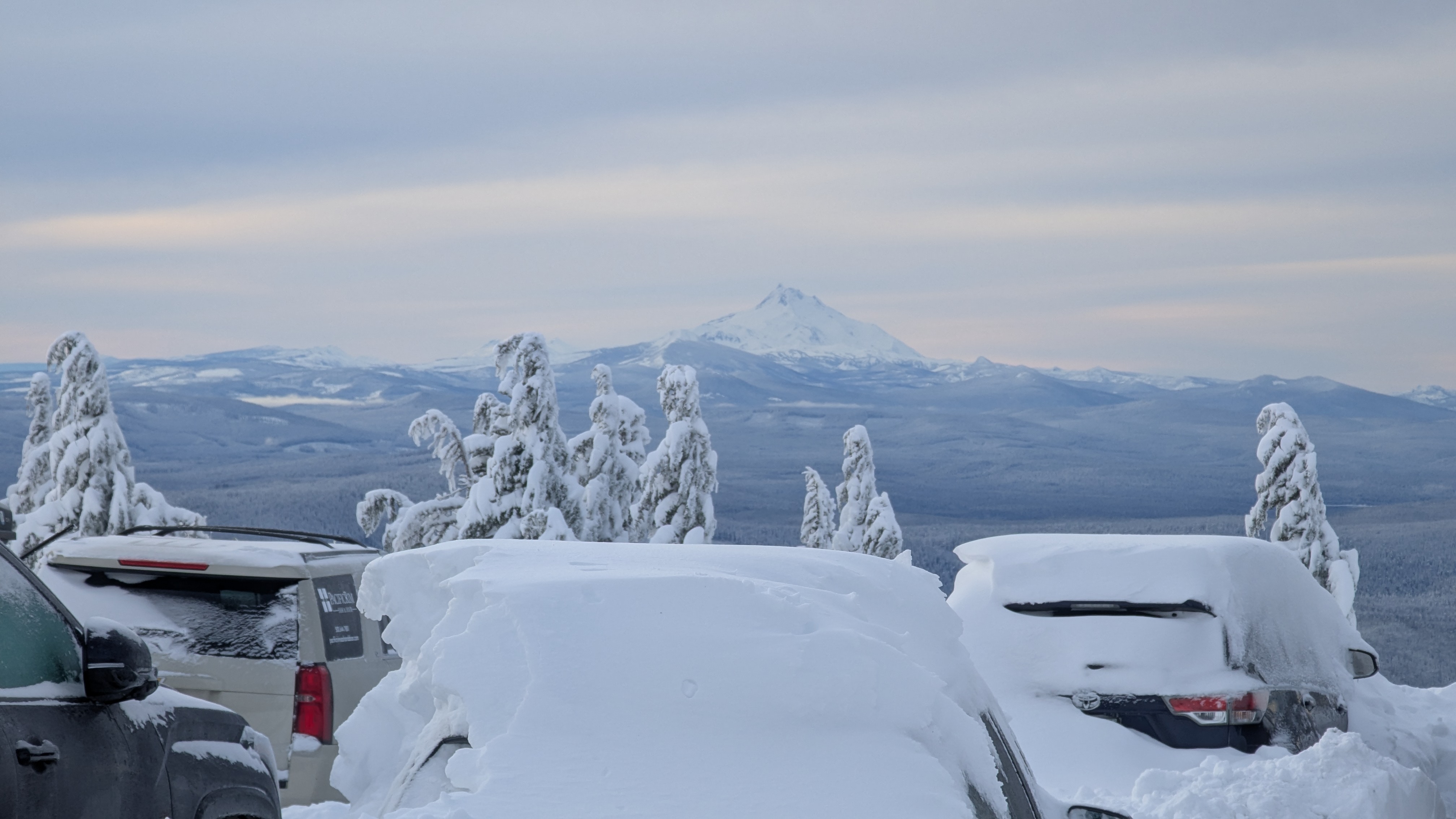 Daytime. Snowy mountain peak of Mt. Jefferson from Timberline's main entrance. The sky is white and steel blue. Cars are packed under a foot of snow. Hood from our hotel room. Lots of snow. Couple conifers caked with snow, too.