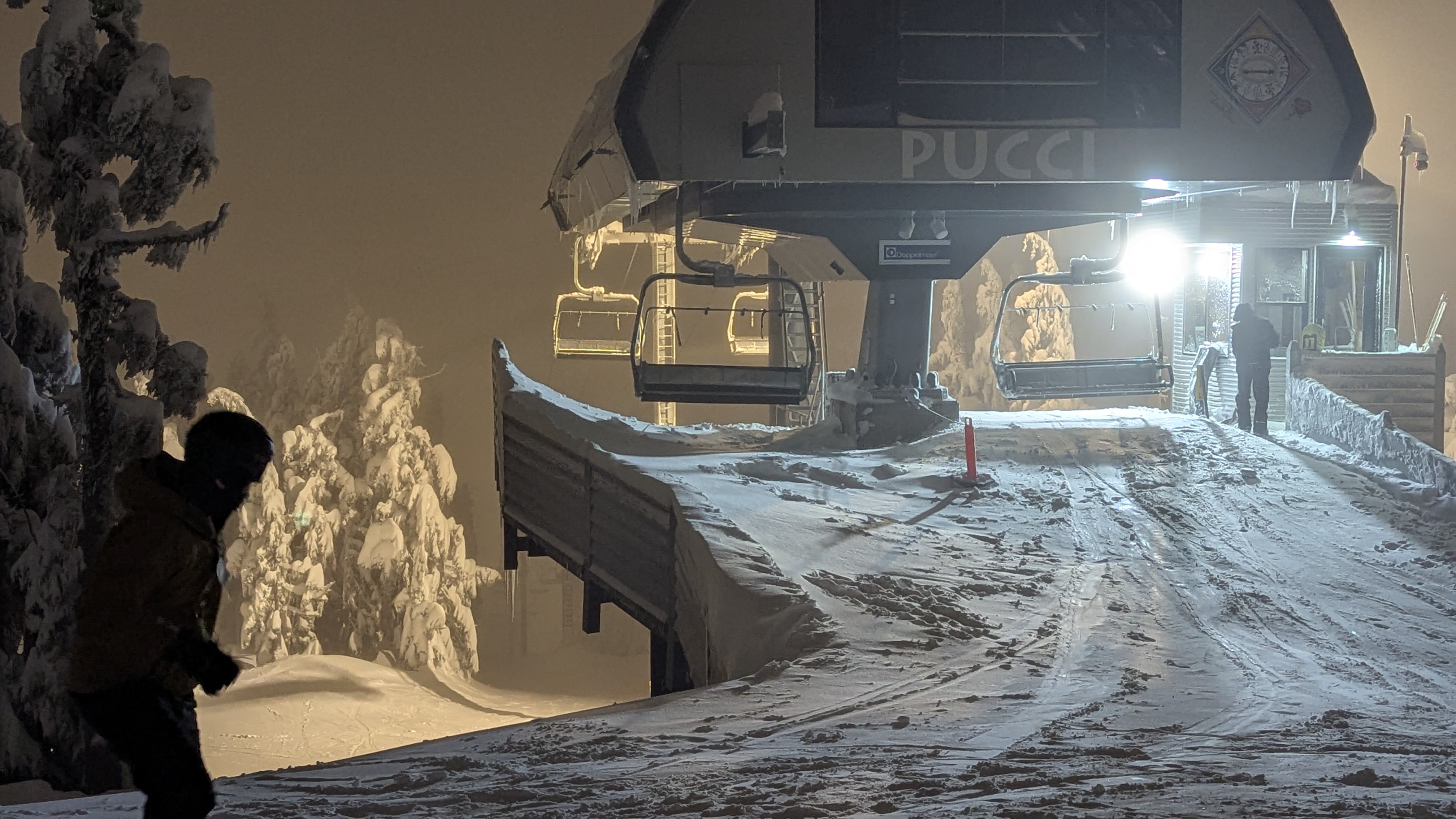Winter alpine setting. Nighttime. From behind, bright lights illuminate a ski lift and operator. Foggy. Snowy. A snowboarder enters the picture from the far left.