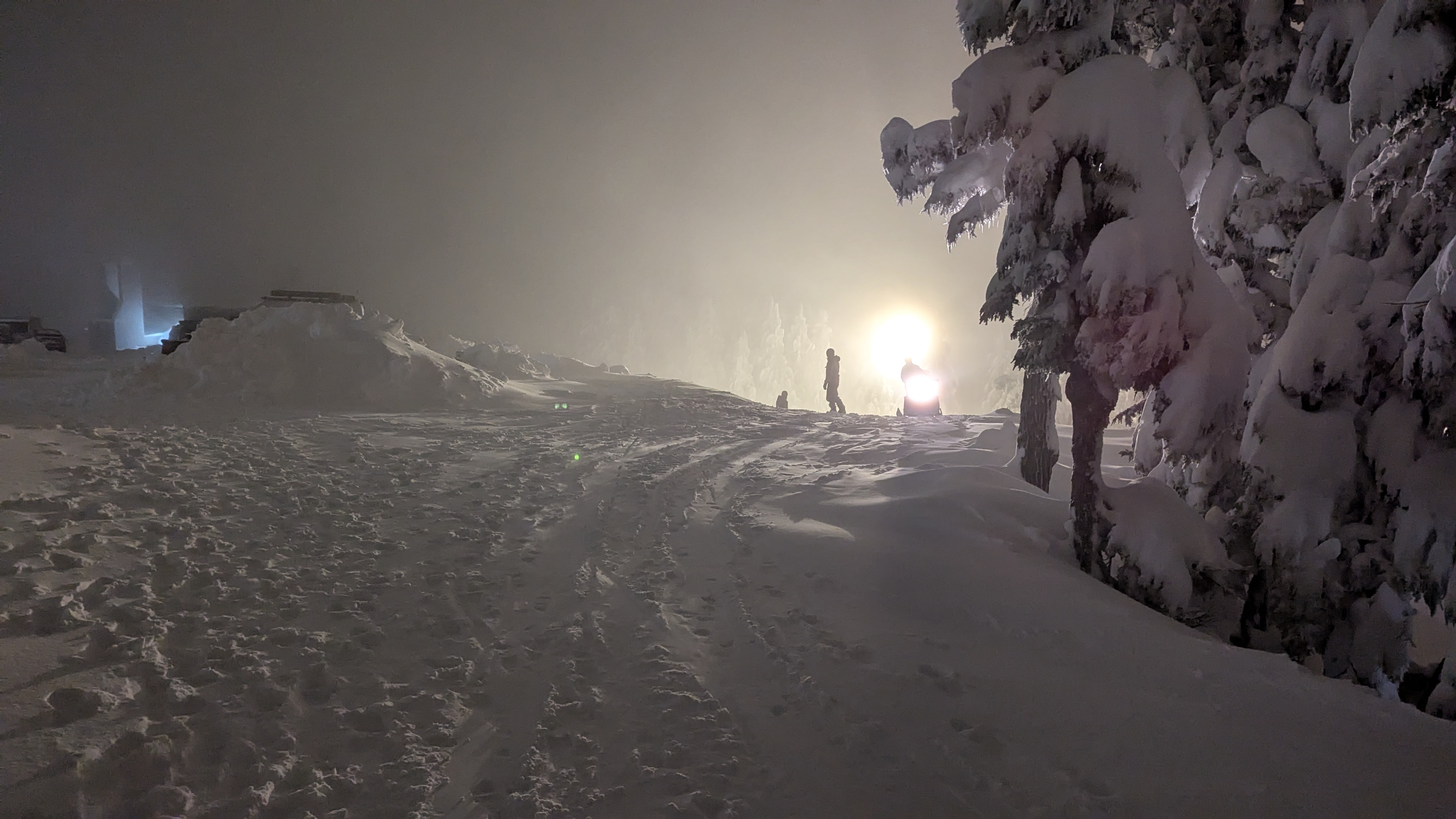 Winter alpine setting. Nighttime. From behind, bright lights illuminate two snowboarders on the crest of a hill starting their run and a ski patrol member on a snowmobile coming up the crest of the hill. Foggy. Snowy.