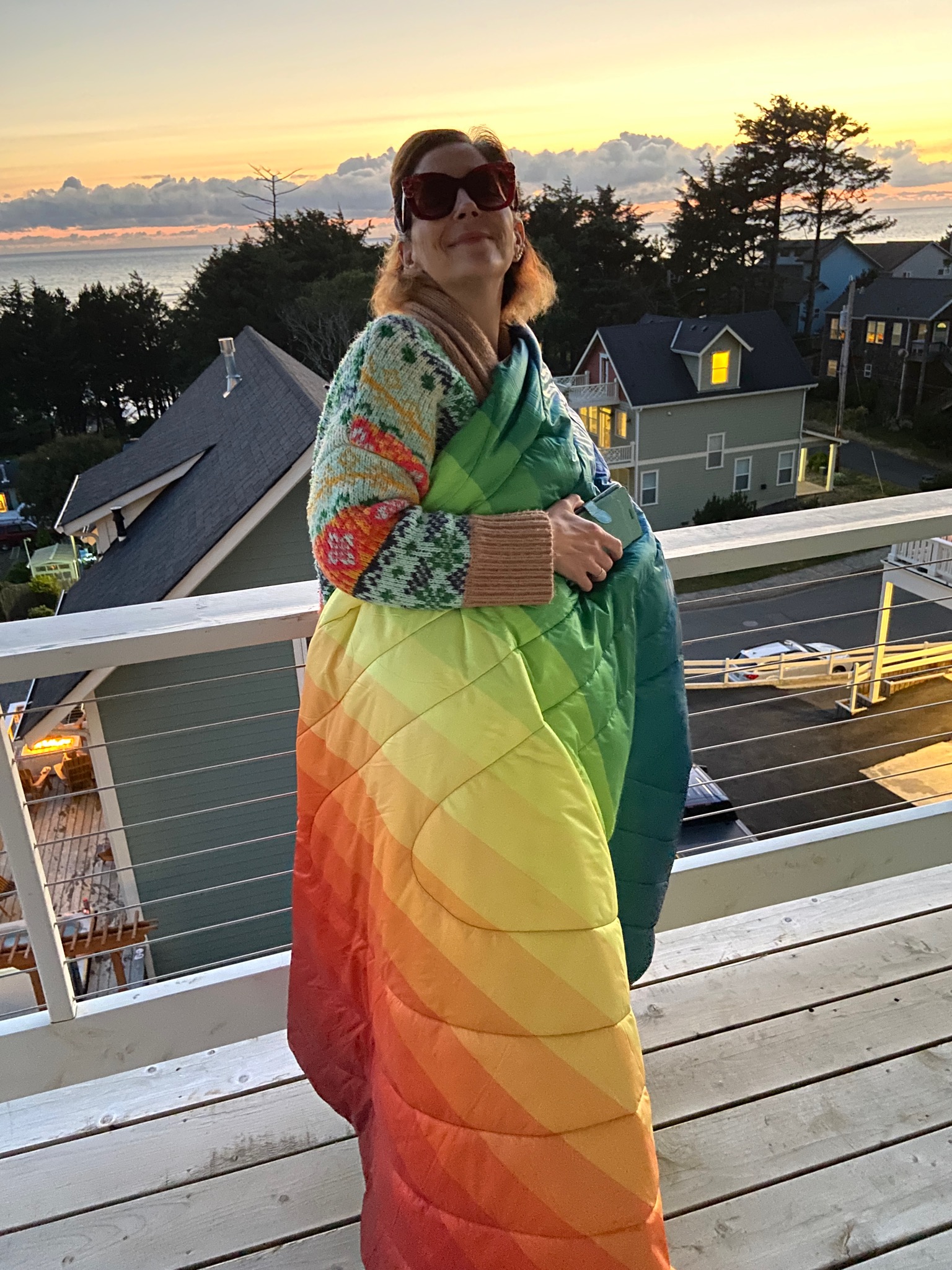 Wife wearing oversized red flower sunglasses wrapped up in a rainbow blanket over a rainbow sweater. She's standing on a deck and the sun is setting behind her over the Pacific Ocean.