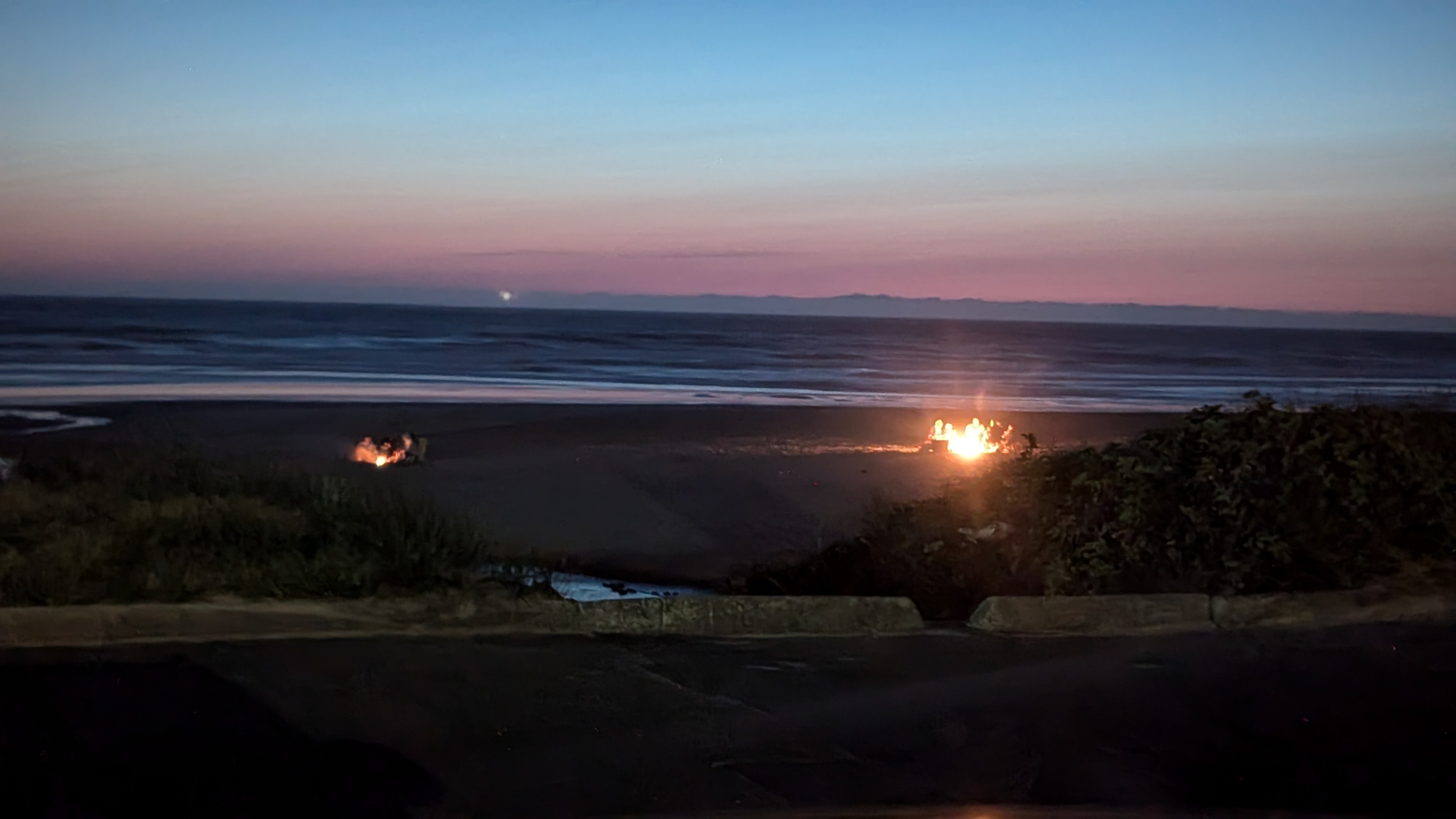 Dusk at a beach on the Pacific Ocean on the Oregon coast. Two bonfires surrounded by people.